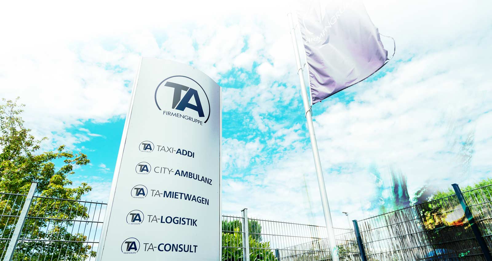 Firmenschild der TA-Firmengruppe mit einer Fahne im Hintergrund